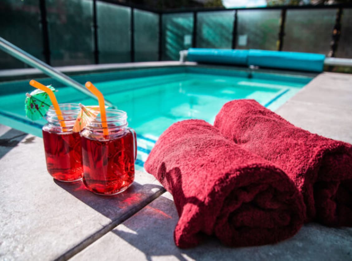 Two mason jars filled with vibrant red beverages are placed on a stone surface alongside two plush red towels. A turquoise pool is visible in the background, surrounded by safety glass fencing, inviting relaxation and enjoyment in a tranquil outdoor setting.