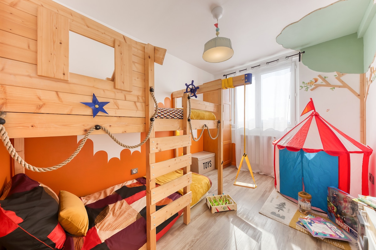 A children's bedroom features a wooden bunk bed with a cozy lower sleeping area and bright bedding. A small circus tent adds a playful touch, while wall decorations showcase vibrant colors. Natural light filters through a nearby window, illuminating the inviting space.