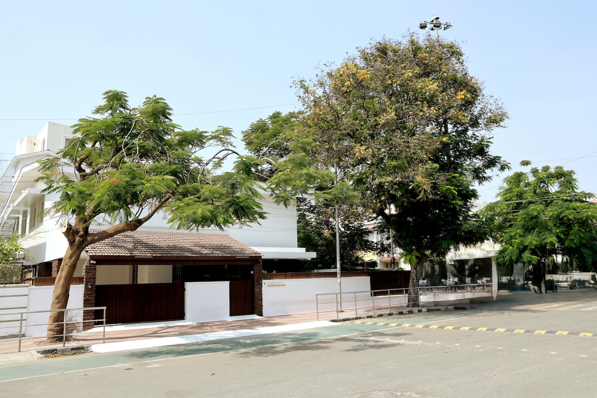 The exterior view of the property displays a single-story structure with a brown wooden gate and a large tree providing shade. Surrounding greenery enhances the inviting atmosphere, while a pedestrian pathway runs along the front, leading towards the building.