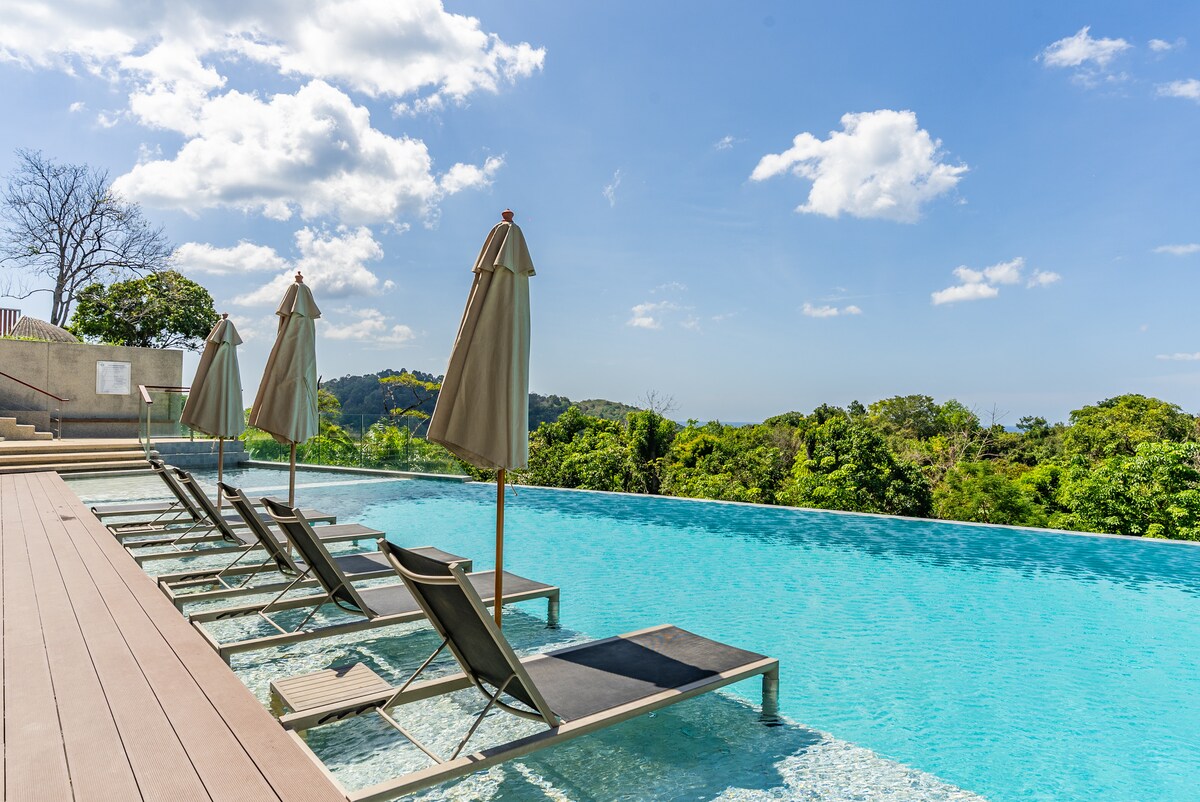 An infinity pool extends towards vibrant greenery, featuring several lounge chairs with umbrellas positioned along the water's edge. The serene environment is complemented by a clear blue sky and soft, scattered clouds, inviting relaxation and cooling off after a warm day.