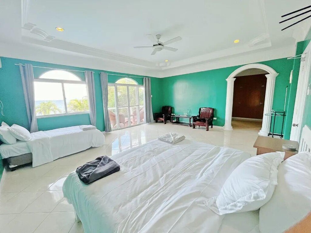 A spacious room features turquoise walls and large windows allowing natural light. Two beds, one with a white coverlet and the other neatly arranged, offer comfortable sleeping arrangements. Two armchairs are positioned nearby, and a doorway leads to another area.