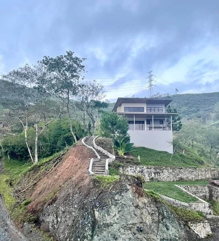 A modern house is positioned on a hillside, surrounded by greenery. A winding stone staircase leads from the lower ground to the spacious balcony above, offering views of the surrounding landscape. The sky is partially cloudy, enhancing the serene mountain atmosphere.