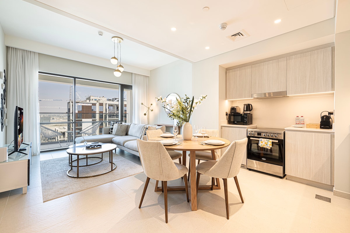 A spacious living area features a comfortable sofa and a smart TV, providing a relaxing space. A dining table with two chairs is set near an open kitchen equipped with modern appliances, including a stove and coffee machine. Large windows offer views of the waterfront.