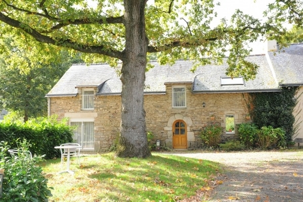 A rustic stone exterior of a two-story building is shown, featuring a welcoming wooden door and large windows. A spacious tree provides shade in the foreground, while well-maintained greenery complements the inviting landscape, creating a peaceful outdoor setting.