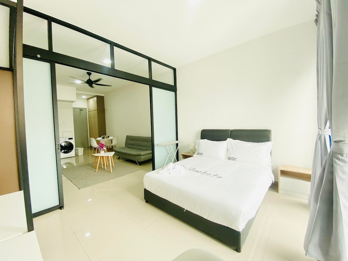 A spacious bedroom features a large bed with crisp linens and decorative pillows. Natural light filters through sheer curtains, illuminating the room. A comfortable seating area is visible to the side, and a modern dining table is positioned in the distance.
