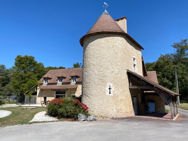 The exterior of a stone and timber gîte is showcased, featuring a tower with a conical roof. A landscaped area with flowers is visible in the foreground, along with a driveway leading to the entrance. Surrounding greenery suggests a peaceful rural setting.