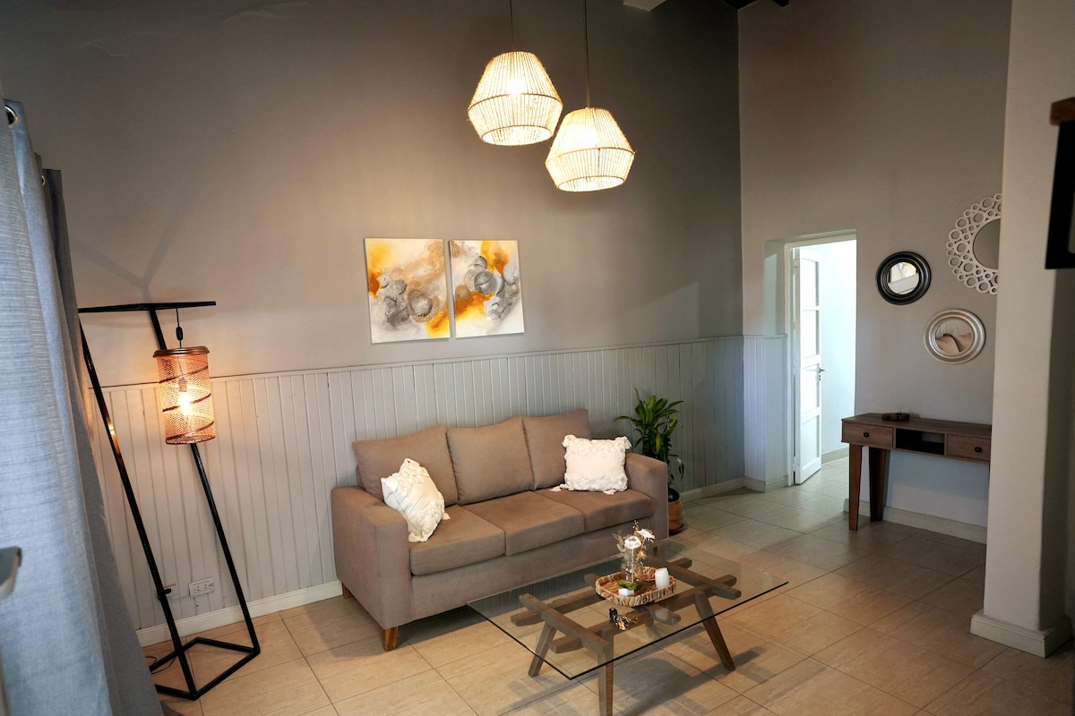 A well-lit living area is highlighted by three pendant lights. A comfortable gray sofa is positioned near a glass coffee table, adorned with decorative items. A subtle accent chair and a small plant add warmth to the space. The room features neutral tones with textured wall accents.