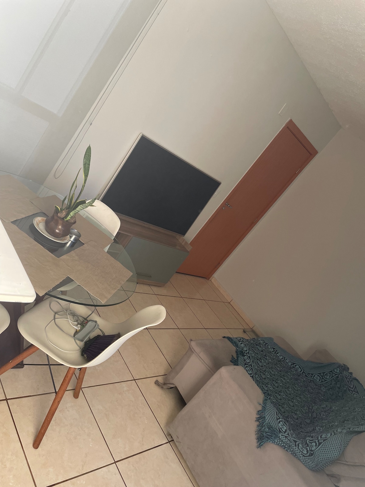 A modern living area is equipped with a glass dining table and white chairs. A smart TV is visible against the wall, with a wooden console beneath. A soft gray sofa is adorned with a decorative throw, providing a relaxed atmosphere. Natural light filters through nearby windows.