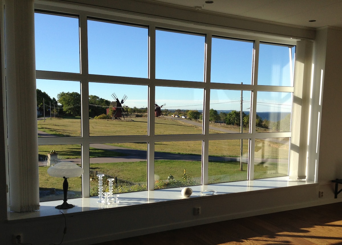 A large window offers an unobstructed view of the open landscape, highlighting traditional red windmills in the distance. Soft natural light filters through the panes, illuminating a simple white lamp and decorative glass pieces on the ledge.