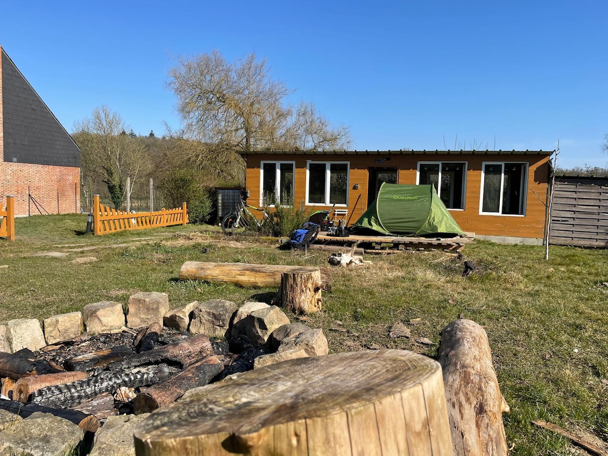 A rustic wooden chalet is nestled in a grassy area, accompanied by a green tent set on a wooden platform. A fire pit made of stones is situated in the foreground, with logs placed around it. Clear skies and greenery frame the peaceful setting.
