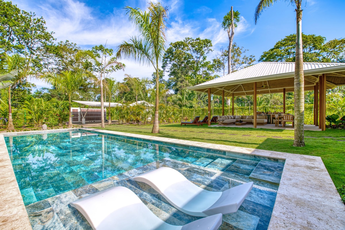 A tranquil outdoor area features a clear swimming pool with two sun loungers partially submerged. Lush greenery surrounds the space, while a covered seating area is visible in the background, equipped with comfortable furnishings. Tall palm trees rise against a blue sky dotted with clouds.