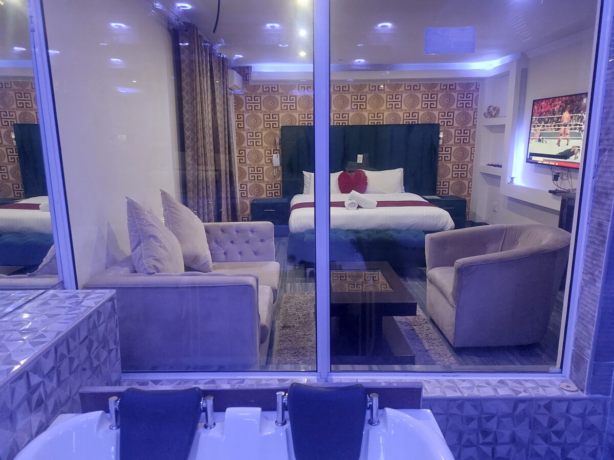 A spacious room features a large bed with decorative pillows, complemented by a seating area with a sofa and two armchairs. The attractive wall design adds character, while a flat-screen TV and modern lighting enhance functionality. A stylish jacuzzi is visible in the foreground.