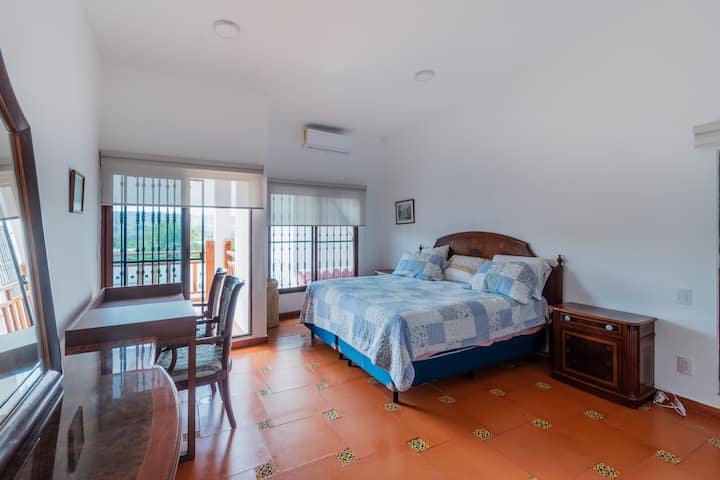 Chambre lumineuse avec lit king size, couvre-lit bleu en patchwork, sol en terre cuite artisanale brillante, bureau avec 2 chaises, climatisation et grandes fenêtres avec balcon et lumière naturelle.