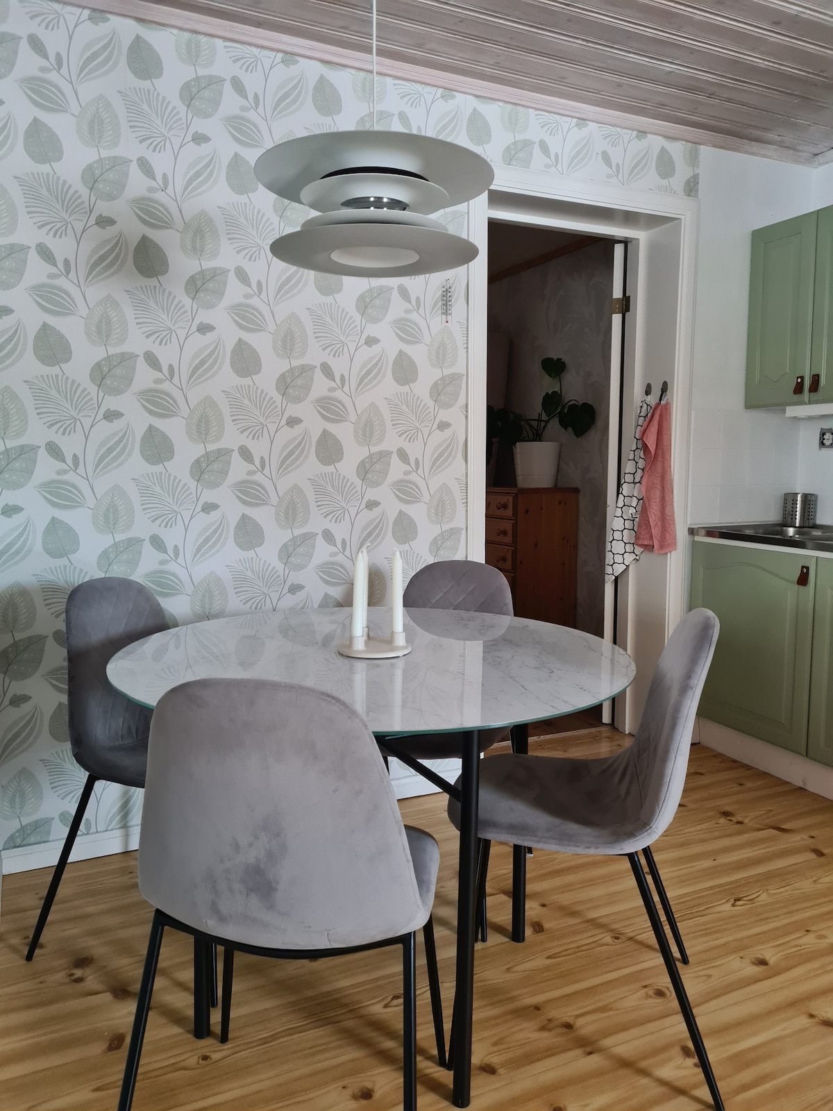 A round dining table with a marble top is surrounded by four upholstered chairs in a soft grey. A modern light fixture hangs above the table, illuminating the space, while a patterned wall with leaf designs adds a natural touch to the room.