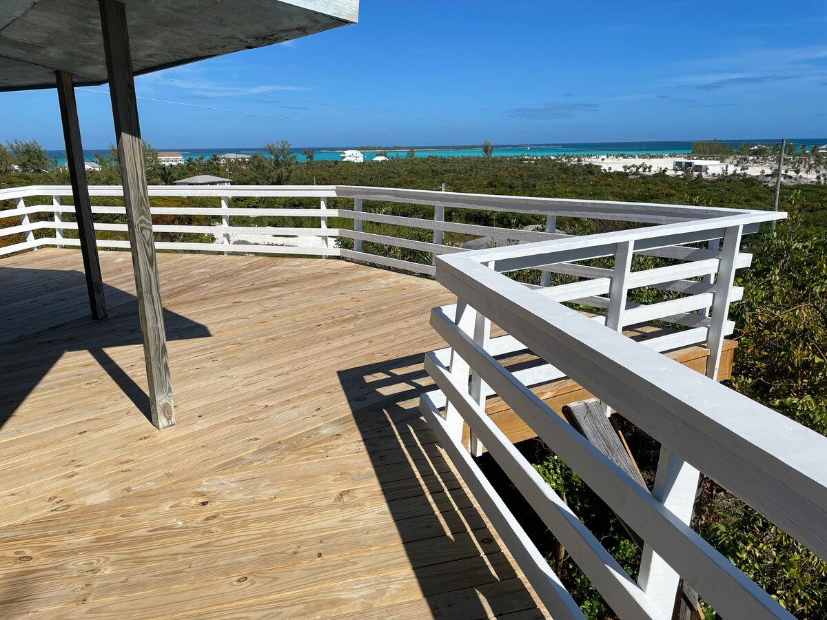 A spacious wooden deck is presented, featuring white railings that frame the horizon view. The turquoise water of the Great Harbour Cay is visible in the distance, with the sandy beach and lush greenery in the foreground under a clear blue sky.