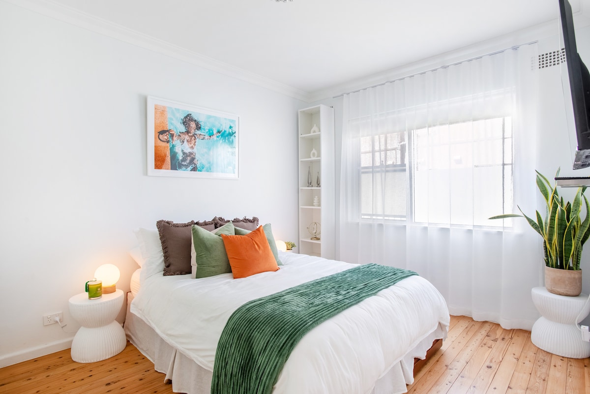 A bright bedroom is presented with a spacious bed topped with assorted pillows, including orange accents. Soft green bedding is showcased alongside light wooden flooring. A window with sheer curtains allows natural light to filter in, and a vibrant artwork piece is displayed on the wall.