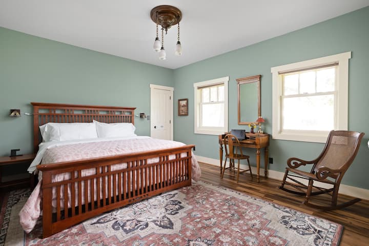 Spacious bedroom featuring a king-sized bed, vintage wood furnishings, a cozy workspace with a desk and chair, and plenty of natural light — perfect for relaxation or remote work.