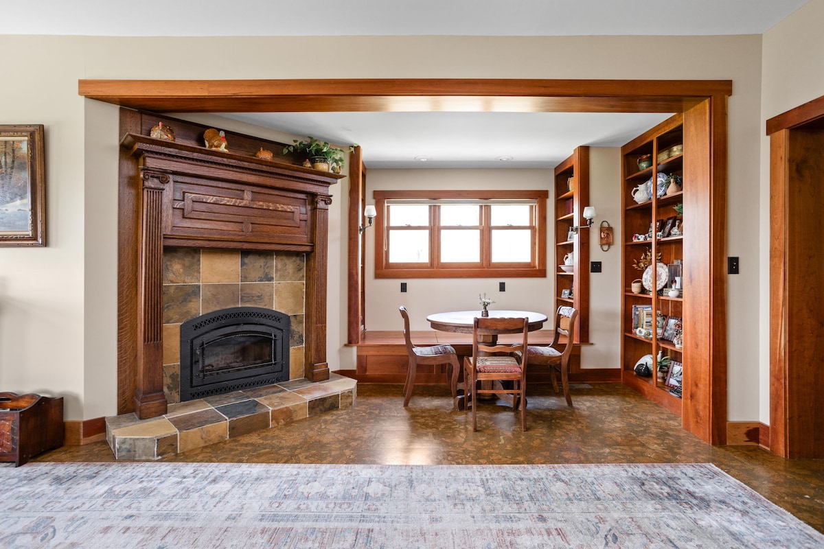 A living room area features a wood-burning fireplace set within a decorative mantle, complemented by built-in bookshelves. A round dining table with four chairs is positioned nearby. Natural light filters in through the large windows, enhancing the warm, wood tones throughout the space.