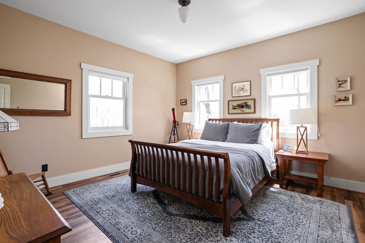 Cozy and inviting bedroom featuring a queen-sized bed, warm natural light, charming artwork, and classic wood furnishings — perfect for a restful stay.