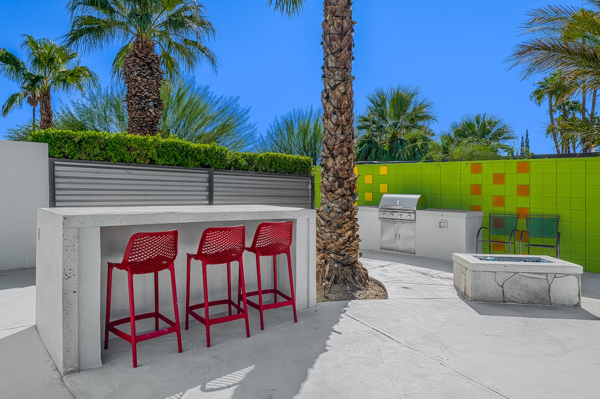 A backyard area is presented with a concrete bar featuring three red stools. Lush palm trees and greenery surround the space, offering privacy. A barbecue grill is positioned nearby, and a fire pit is visible in the corner, contributing to a relaxed outdoor environment.