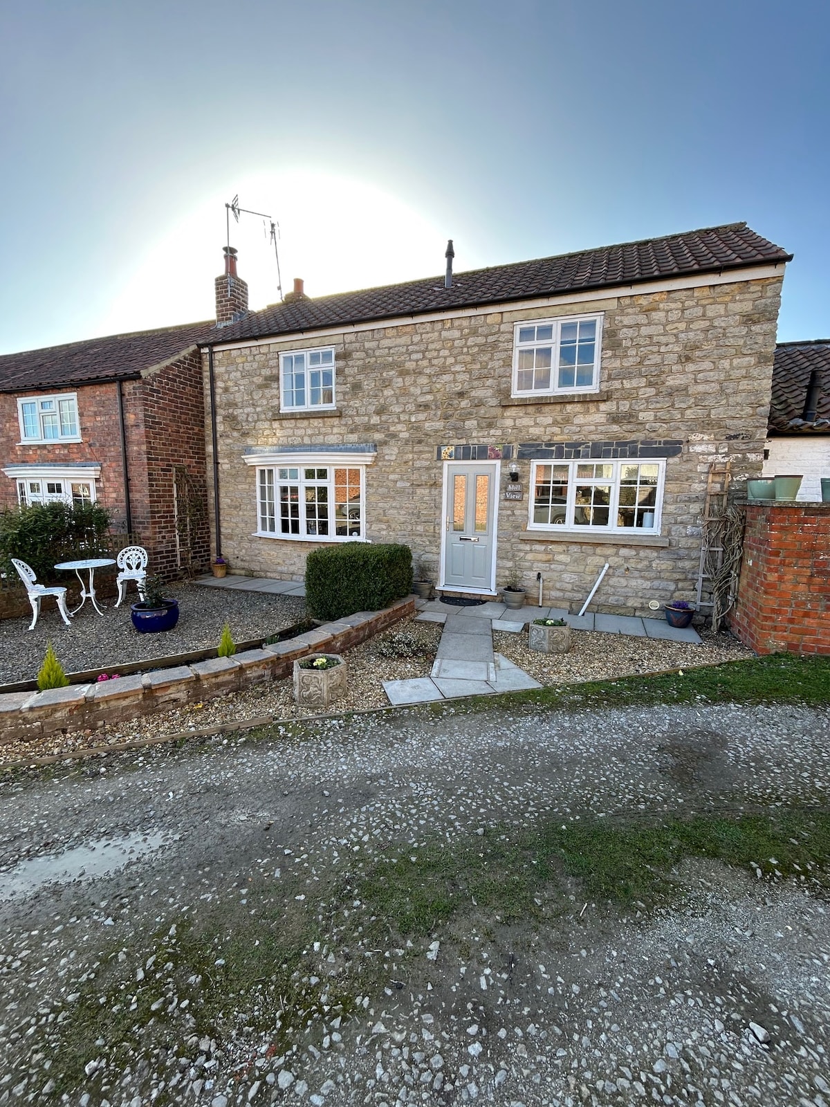 A charming stone cottage is viewed from the front, featuring a light grey door flanked by two windows. A small courtyard with patio seating is visible, surrounded by gravel and bordered by neat greenery. Neighbouring homes provide a quaint backdrop.
