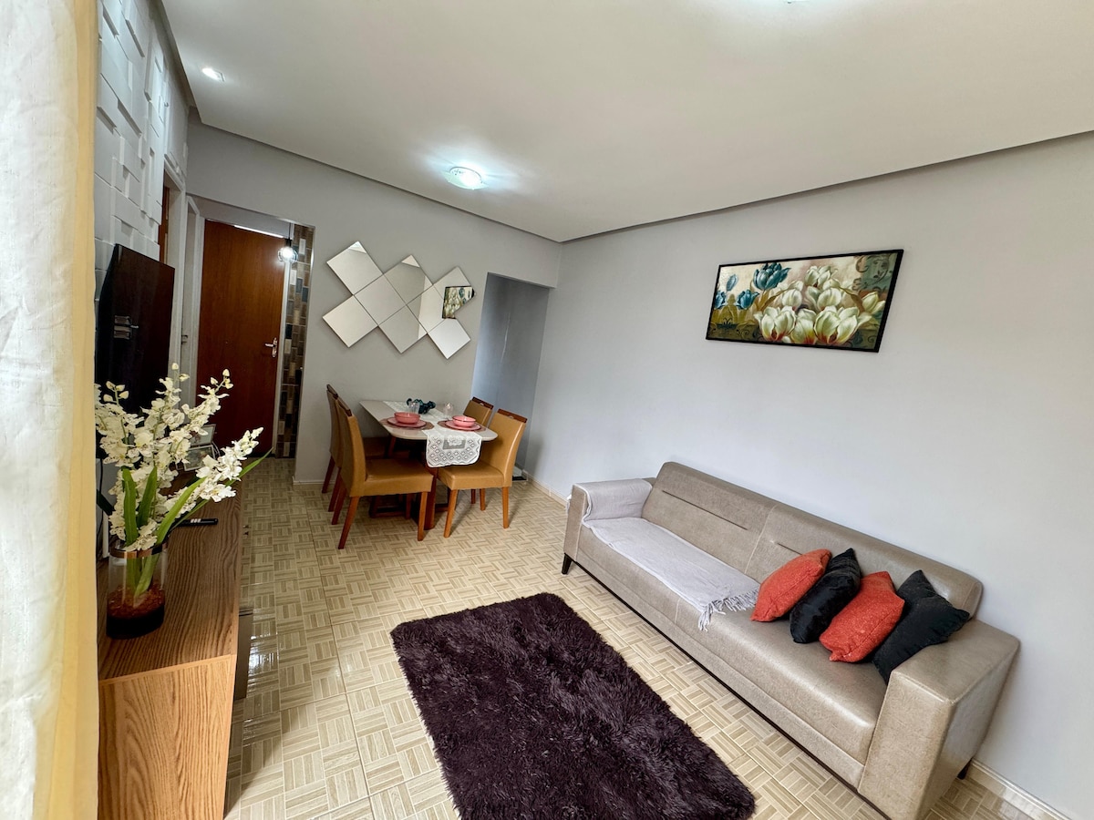 A bright living area features a light-colored sofa with decorative cushions and a soft rug on a patterned floor. A dining table is set for four, and a piece of artwork adorns the wall. Natural light enhances the inviting space.