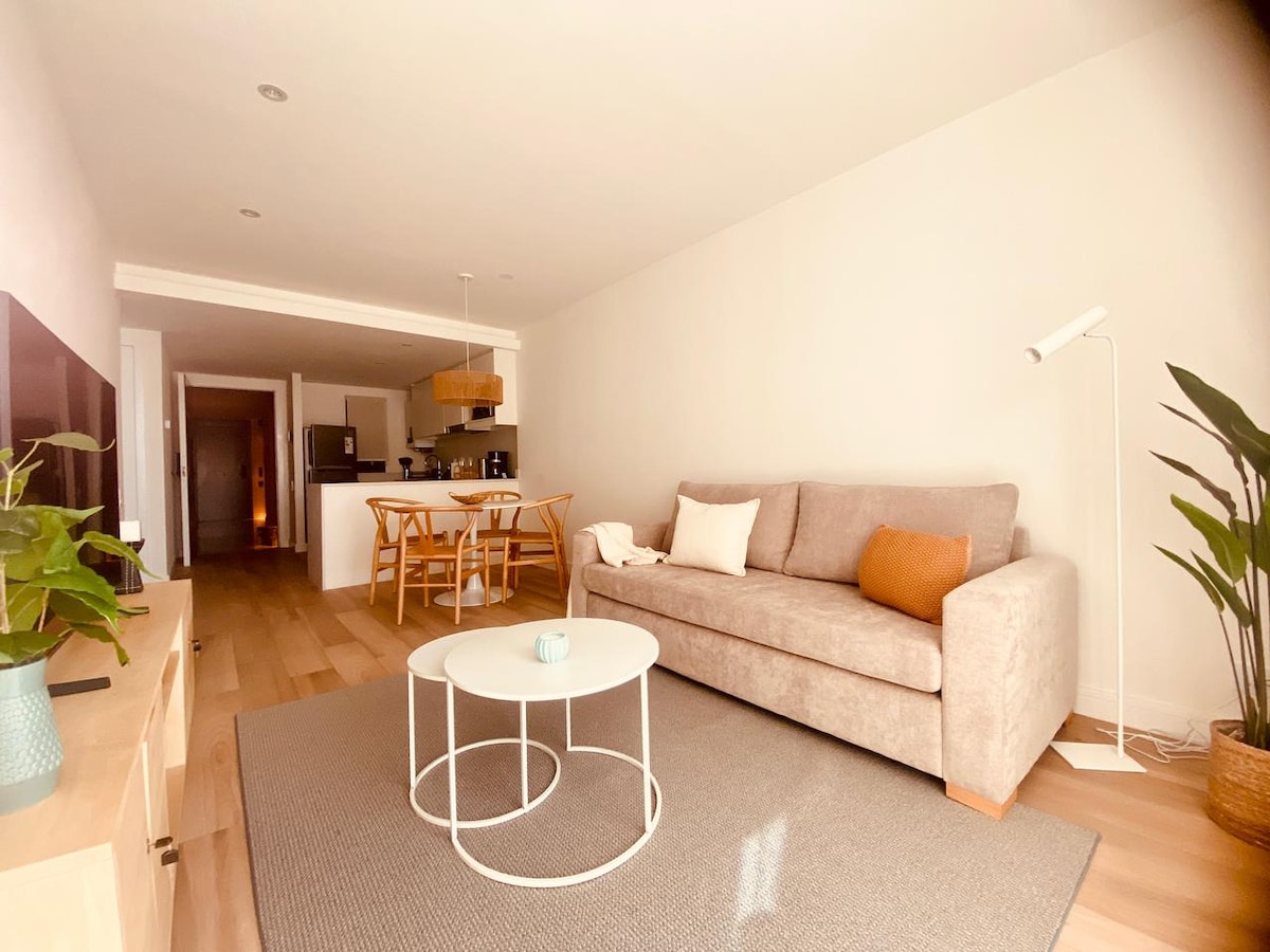 A spacious living area showcases a neutral color palette with natural light illuminating a comfortable sofa and a round coffee table. A dining table with chairs is visible in the semi-integrated kitchen area, complemented by decorative plants positioned nearby.