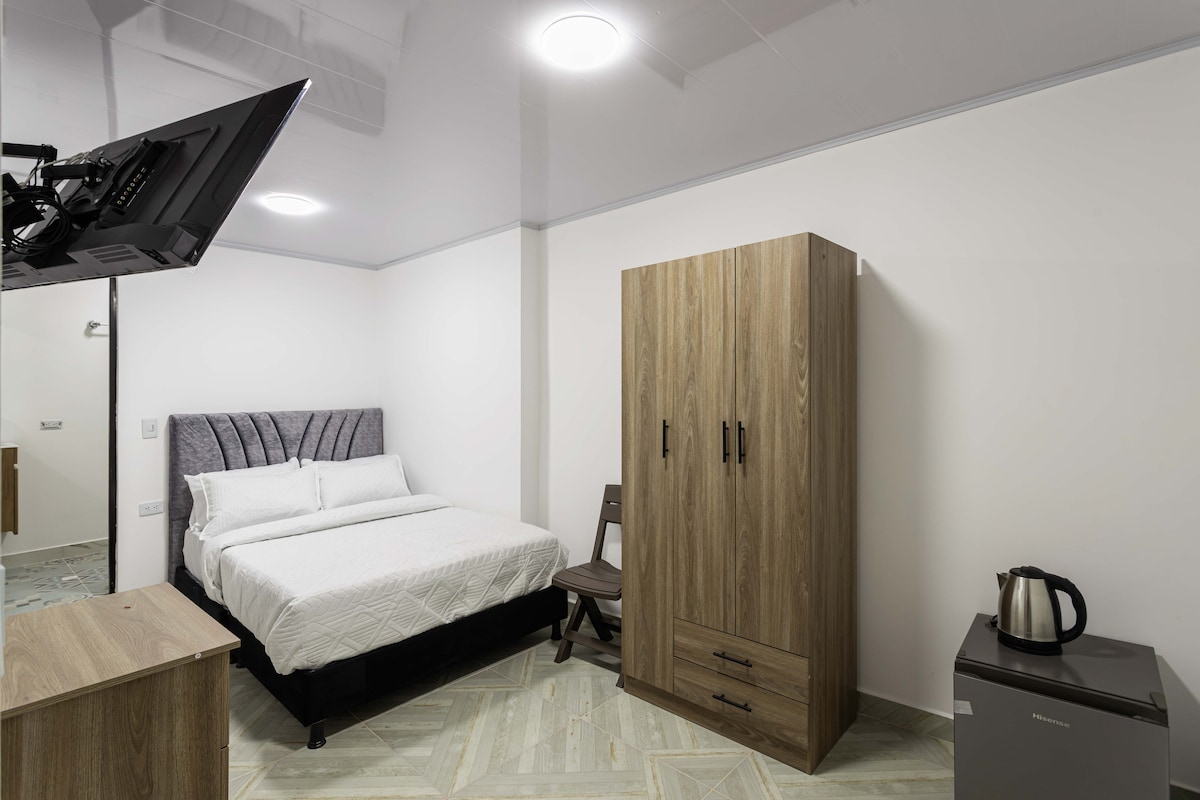 A private room features a comfortable bed with a neutral-colored bedspread. An efficiently arranged wardrobe and a small bedside table complement the space, while a television is mounted on the wall. A kettle sits atop a compact refrigerator, enhancing the room's convenience.