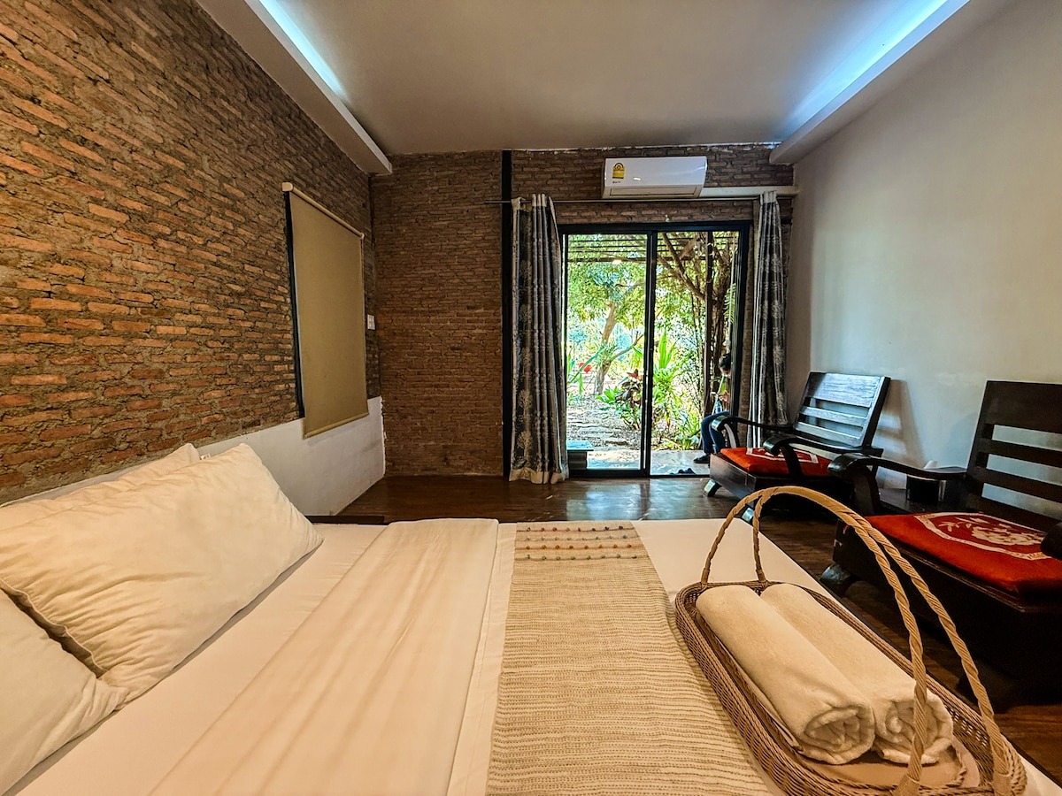 A serene sleeping area features a spacious bed with crisp white linens. Soft natural light filters through glass doors that open to a garden view. Complementary seating is provided with two wooden chairs, and a decorative basket holding rolled towels is placed near the bed.