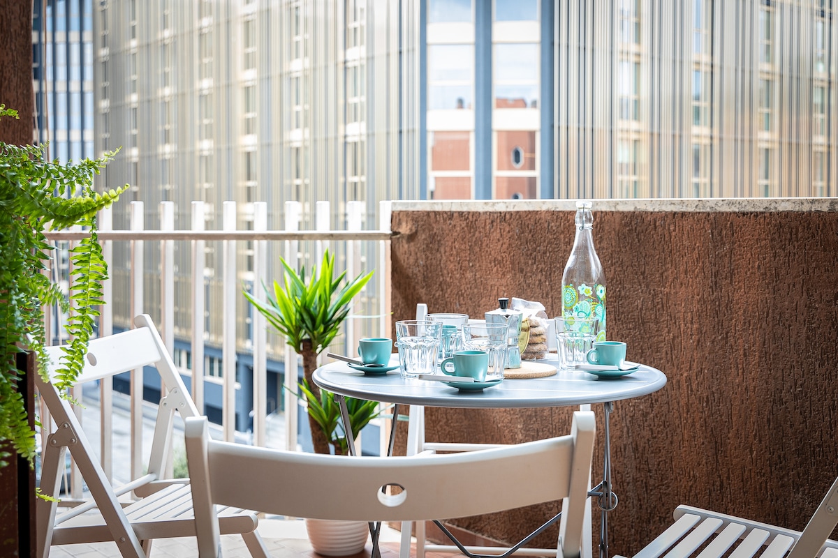 A small outdoor terrace features a round table set for coffee, accompanied by two chairs. A glass water carafe and matching cups are placed on the table, complemented by greenery in the background. The view includes a backdrop of modern building facades.