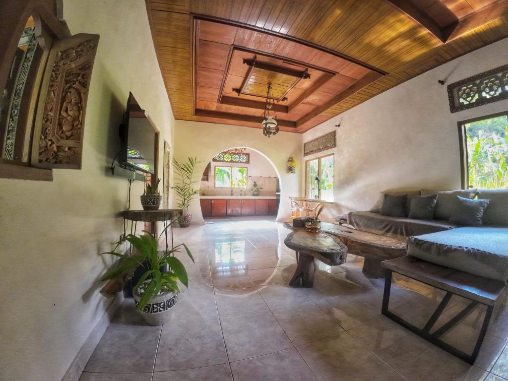 An open-air living area is characterized by a wooden ceiling and intricate wall carvings. A cozy sectional sofa and a low wooden table occupy the space. Large windows provide views of the lush surroundings, allowing natural light to fill the room.