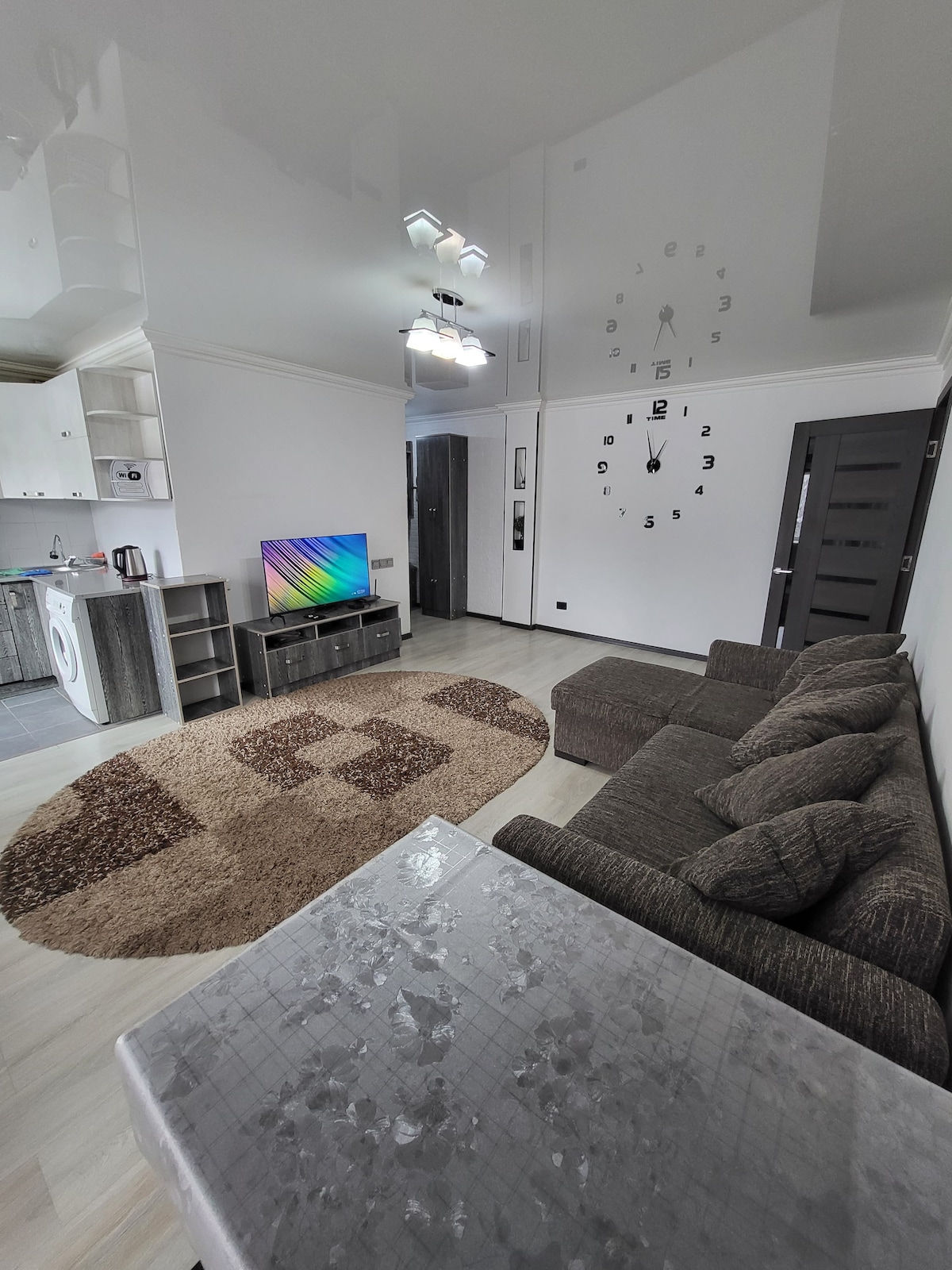 A well-arranged living area features a soft sofa in a neutral tone, positioned near a modern television. An area rug with a subtle design covers the floor, while a reflective table adds a touch of elegance. Bright overhead lighting complements the room's spaciousness.