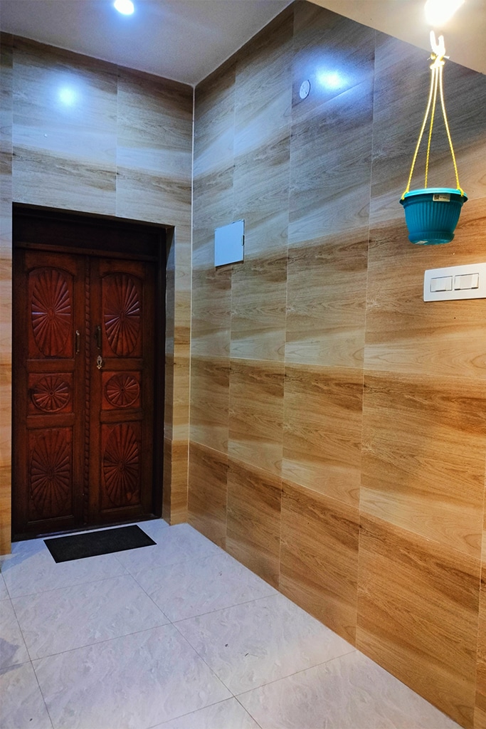 The entrance features a solid wood door with carved detailing, complemented by warm wood paneling on the walls. A small mat lies at the doorway, while a ceiling light illuminates the area. A hanging plant pot adds a touch of greenery.