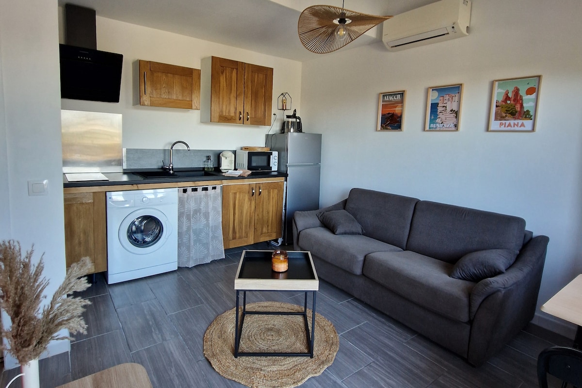 A modern kitchen area features wooden cabinets, a stainless steel sink, and appliances including a microwave and refrigerator. A washer is positioned prominently. A dark gray sofa and a round coffee table with a candle are placed on a woven rug, adding comfort to the space.