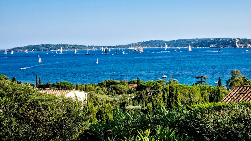 A sweeping view of the azure sea is presented, filled with numerous sailboats gently gliding across the water. Verdant hills are framed by lush greenery, enhancing the serene coastal landscape leading to the distant shoreline.