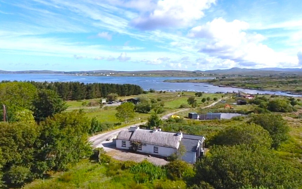 An aerial view showcases the thatched cottage nestled in lush greenery, with the serene Traighenna Bay visible in the distance. Expansive blue skies dotted with clouds add to the peaceful setting. The surrounding countryside includes trees and open fields, enhancing the tranquil atmosphere.