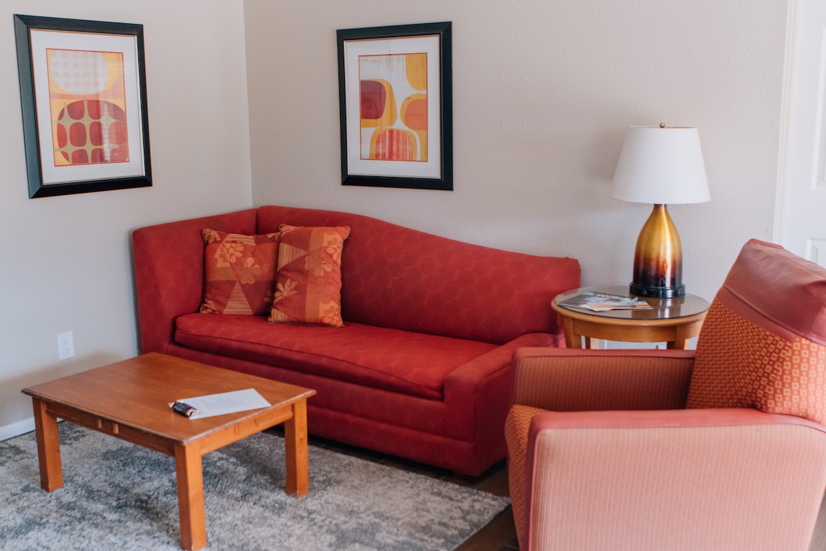 A cozy living area is furnished with an elongated red sofa adorned with patterned cushions, accompanied by an orange accent chair. A wooden coffee table sits centrally, supporting a notepad. Two framed artworks hang on the wall, adding subtle color to the space.