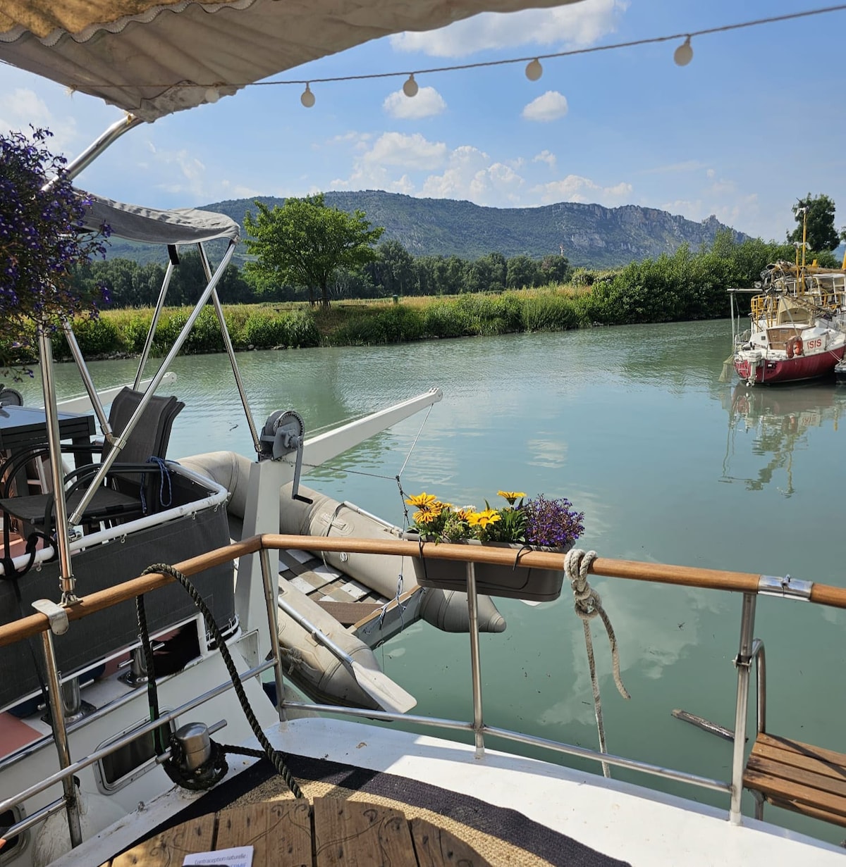 A scenic view from the boat, featuring a calm waterway bordered by lush greenery and distant hills. Flowers are planted in a container, adding color to the space. Nearby, another boat is anchored, contributing to the serene atmosphere.