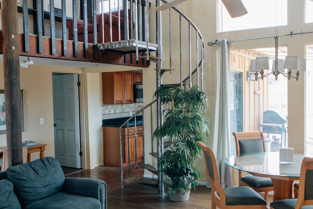 An open living area features a comfortable seating arrangement and a dining table. A spiral staircase leads to a loft with additional sleeping space. Natural light pours in through large windows, highlighting the stylish kitchen in the background, and a greenery accent adds life to the space.