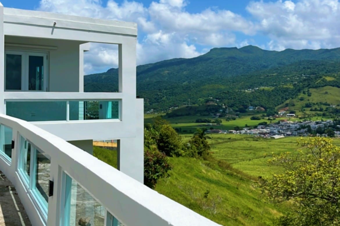 A modern building features a sleek balcony with glass railings, providing unobstructed views of lush green hills and mountains in the background. The sky above is dotted with fluffy clouds, enhancing the sense of tranquility in the natural landscape.