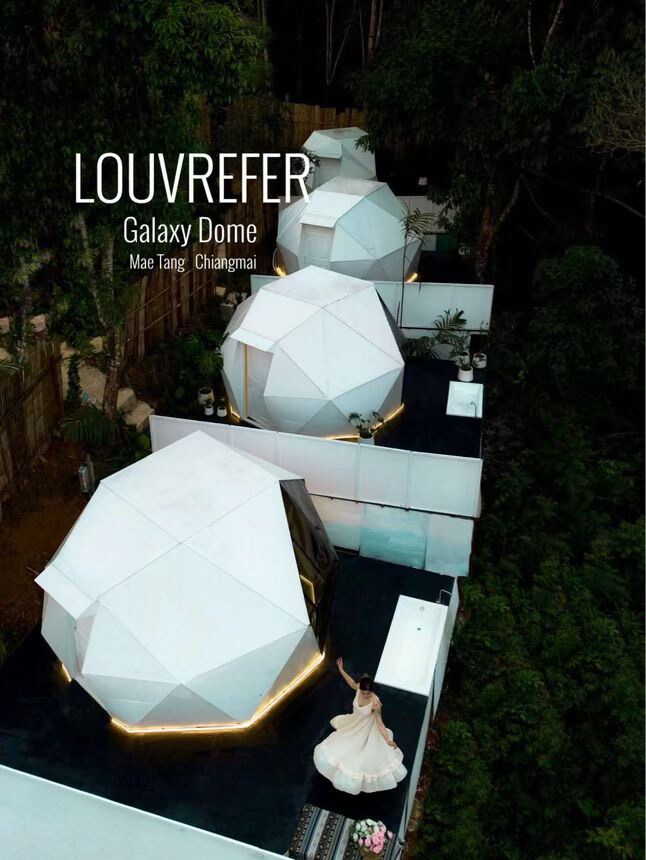 Aerial view of several geodesic domes surrounded by lush greenery. The domes are uniquely designed with white panels, featuring soft lighting along their edges. A pathway connects the domes, providing an inviting atmosphere for relaxation and exploration.