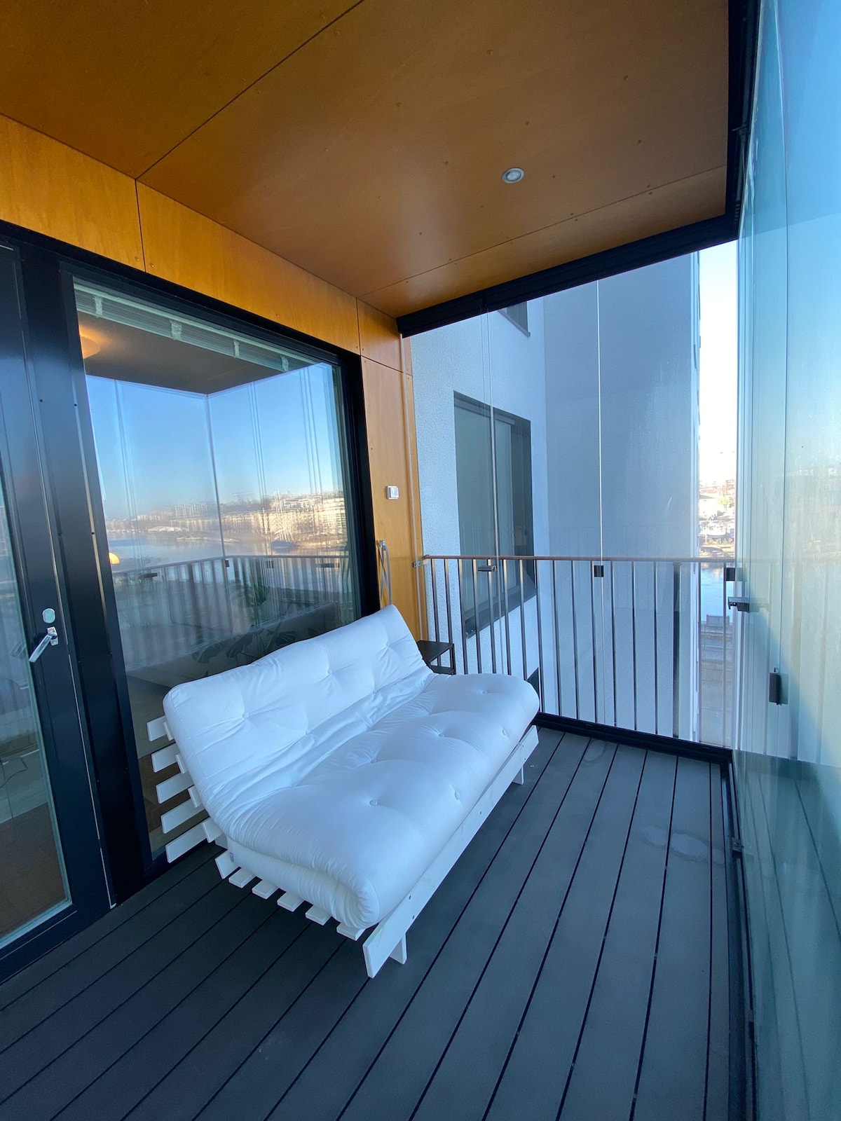 A seating area is present on a glass-enclosed terrace, featuring a white sofa against a backdrop of large windows. The flooring is made of dark wood, and views of the surroundings are visible through the transparent walls.