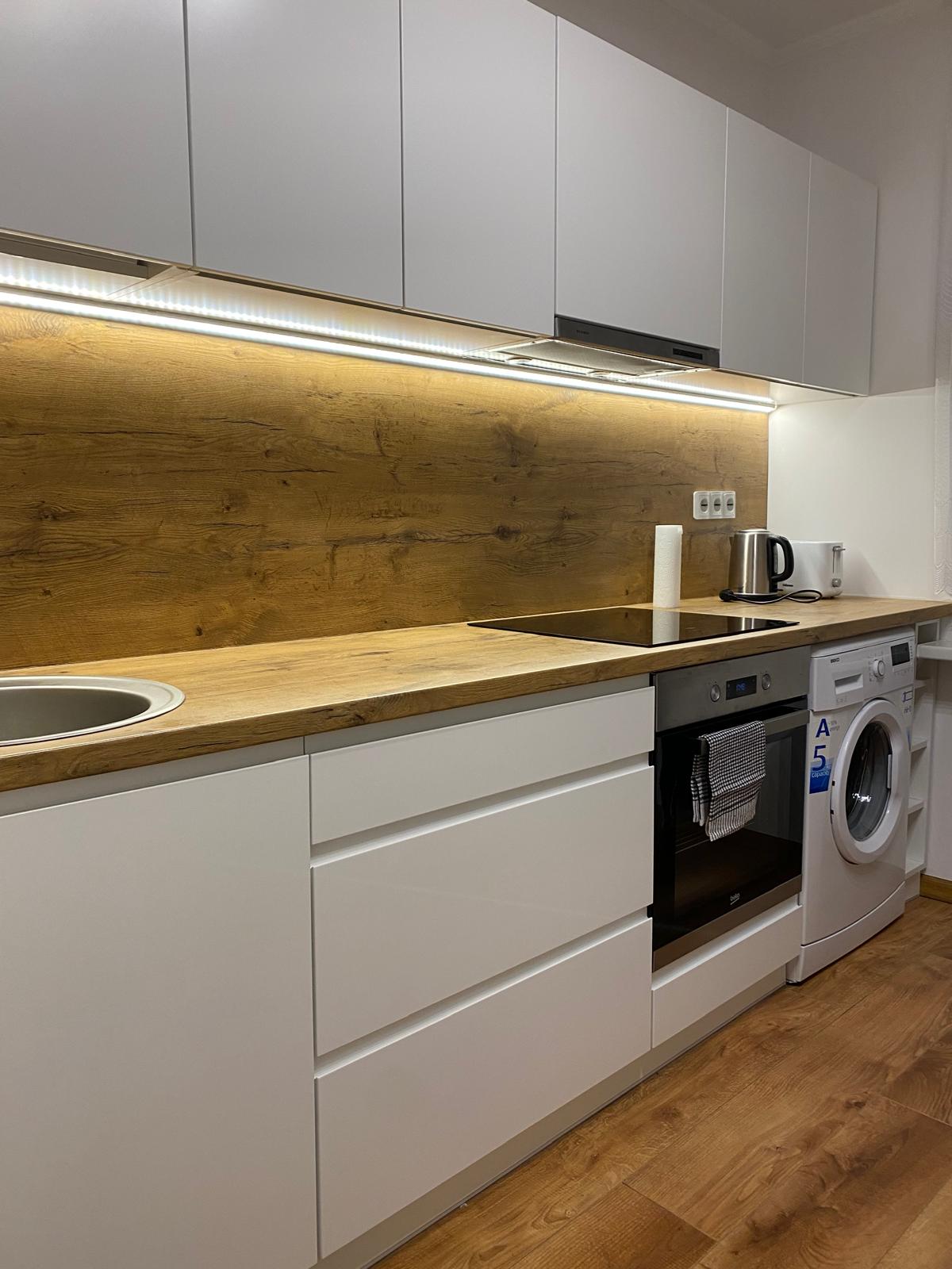 A modern kitchen features sleek white cabinetry and a warm wooden backsplash. An integrated stove and oven are positioned beneath a kitchen hood. A washing machine sits adjacent to the countertop. Soft lighting highlights the cooking area, creating a functional and inviting space.