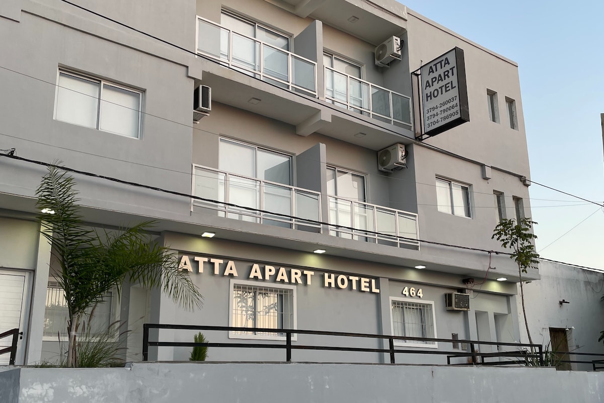 The exterior of Atta Apart Hotel is presented, featuring a modern grey façade with large windows and balconies. The hotel name is prominently displayed on a sign. A small palm plant is positioned at the entrance, adding a touch of greenery.