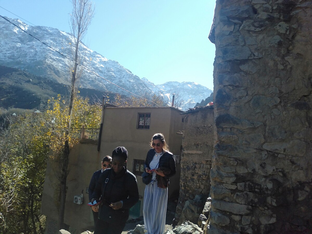 Two people are seen walking near a rustic stone building. Behind them, snow-capped mountains rise against a clear blue sky, and trees with autumn foliage are visible on the left side of the image.