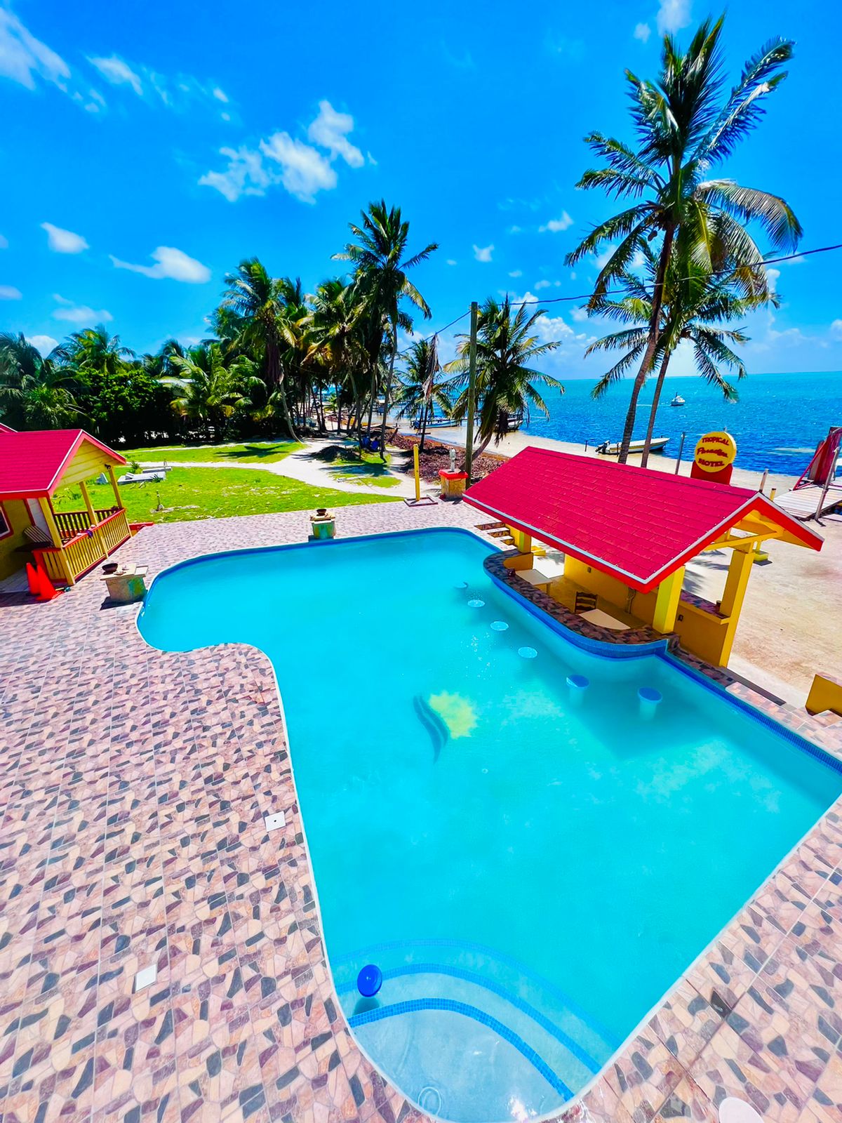 A large swimming pool is surrounded by a vibrant pool deck, featuring colorful cabanas and palm trees. The clear blue water reflects the sunny sky, while the beach area is visible in the background, providing an inviting space for relaxation.
