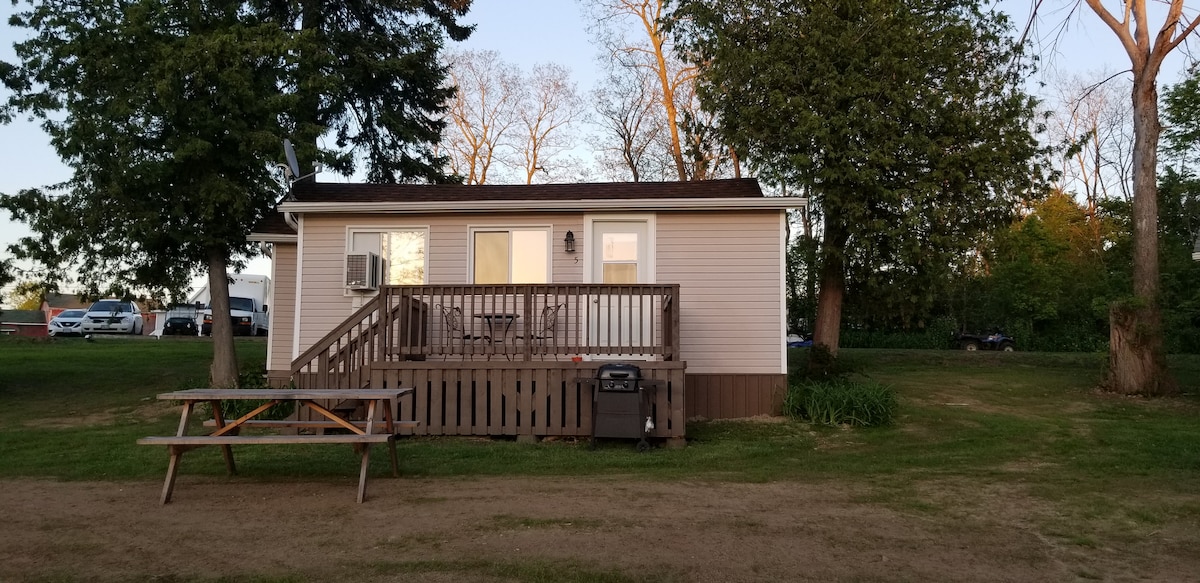 A simple cottage is presented with light-colored siding and a welcoming porch. A wooden picnic table is positioned on the lawn, with a grill nearby. Light from the windows suggests a cozy interior, while trees outline the scene in the background.