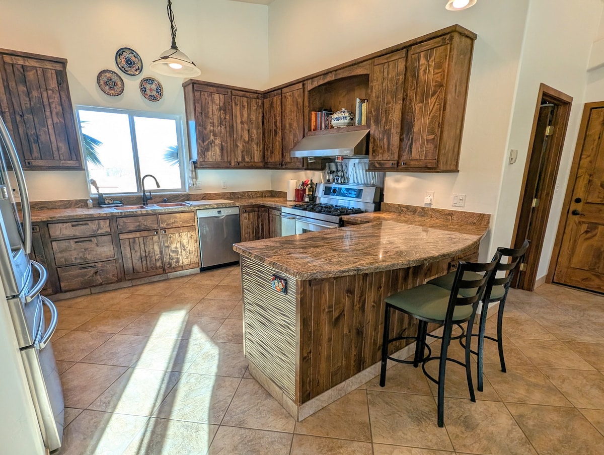The kitchen features custom wooden cabinetry and granite countertops. A spacious island with two bar stools provides additional seating. Stainless steel appliances, including a dishwasher and gas range, are visible, along with a window offering natural light and views of the surrounding landscape.