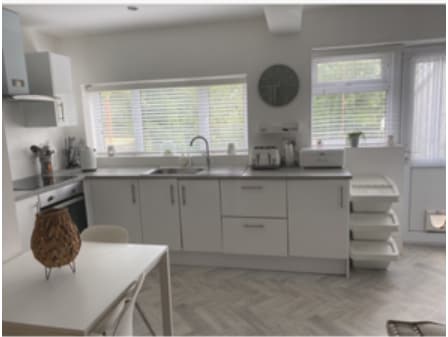 A modern kitchen space is presented with white cabinetry and a neutral countertop. Natural light brightens the area through multiple windows. Essential appliances, including an oven and kettle, are neatly arranged. A small dining table is positioned nearby, complemented by light-colored chairs.