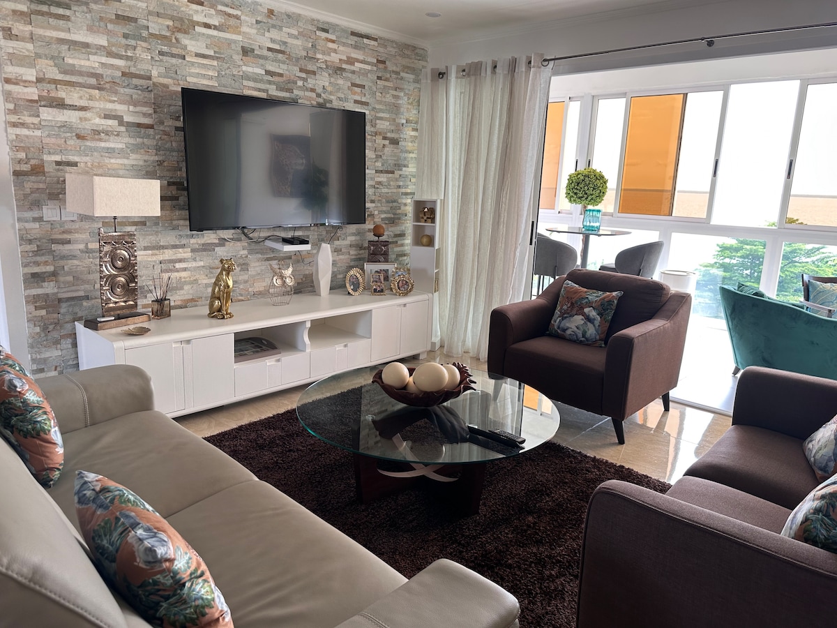 A modern living area features a neutral color palette with a stone accent wall. A comfortable sectional and a brown armchair are arranged around a glass coffee table. Natural light enters through large windows, illuminating decorative items on a white media console.